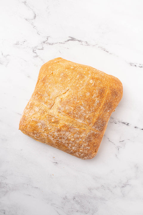 A rustic rectangular loaf of bread with a golden-brown crust and a light dusting of flour, placed on a white marble surface.