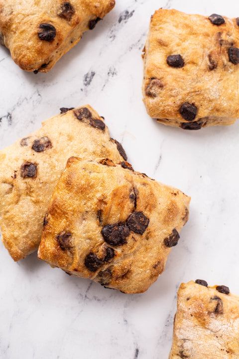 Six baked chocolate chip mini ciabatta with golden-brown crusts and dark chocolate chips, arranged on a white marble surface.