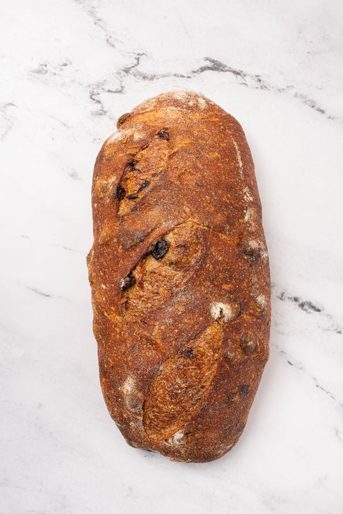 Close up artisanal sourdough raisin bread loaf with a golden-brown crust and dark charred scoring, placed on a white marble surface.