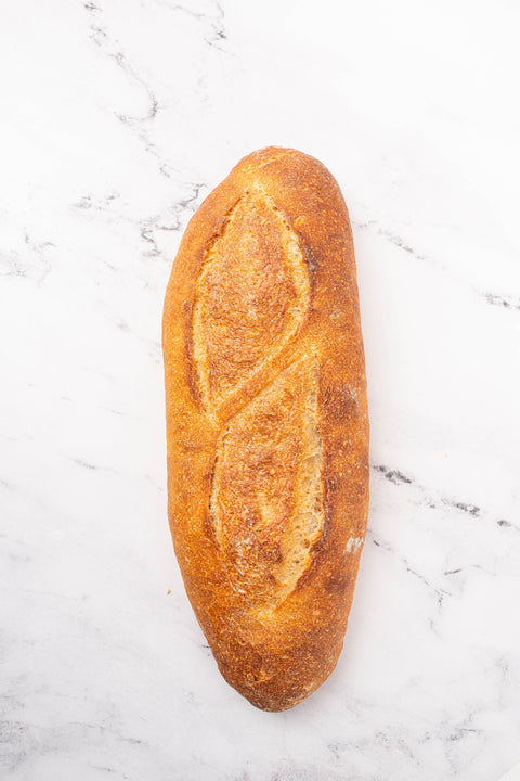 A single rustic loaf of bread with a golden-brown crust and decorative scoring, placed on a white marble surface.