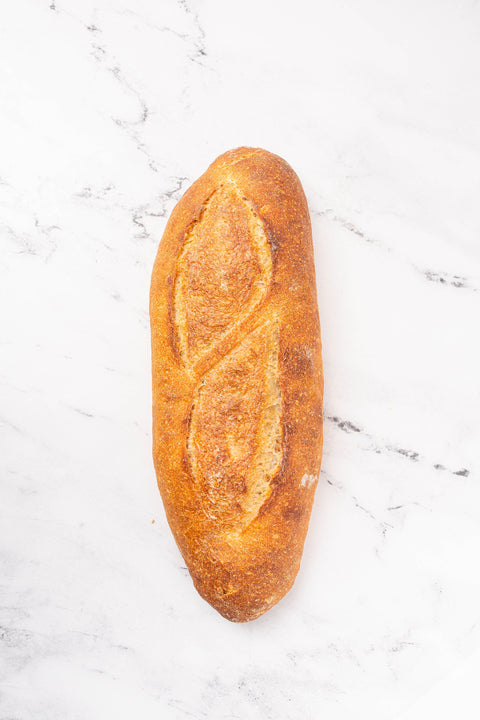 A single rustic artisanal baguette with a golden-brown crust and decorative scoring, placed vertically on a white marble surface with gray veining.