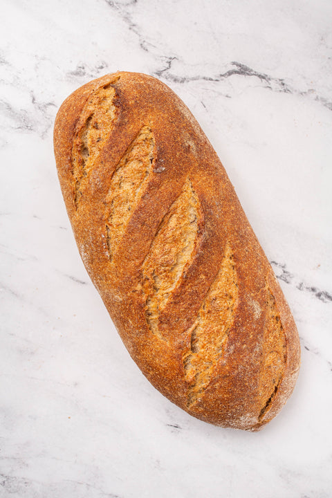 A single rustic seeded rye loaf of bread with a golden-brown crust and scored slashes, placed diagonally on a white marble surface.