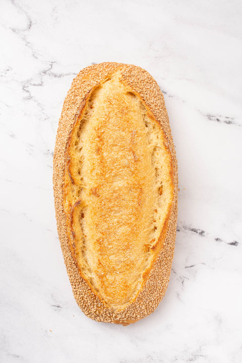 An oval baguette with a golden-brown crust and sesame seeds, photographed from above on a white marble surface.