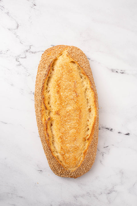 An oval semolina baguette with a golden-brown crust and sesame seeds, photographed from above on a white marble surface.