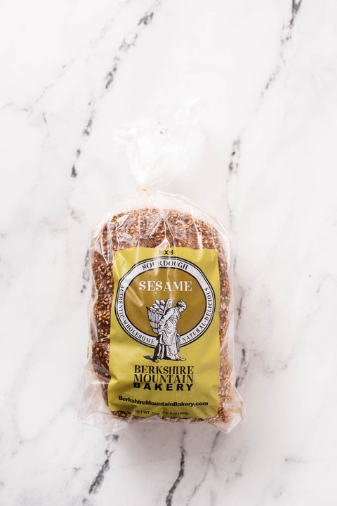 A loaf of sesame seed bread in a clear plastic bag with a yellow label reading 'Sesame' and 'BerkshireMountain Bakery,' placed on a white marble surface.