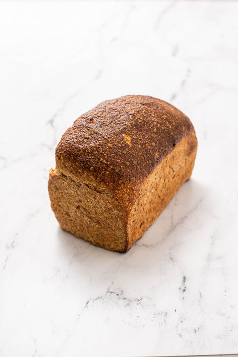 A single loaf of rustic spelt bread sits on a white marble surface with gray veining.
