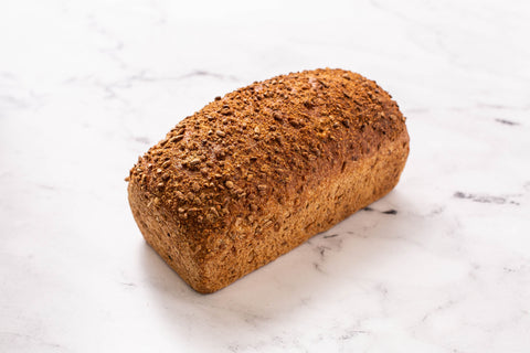 A single loaf of rustic whole wheat bread with a textured, seeded flax crust, photographed on a white marble surface.