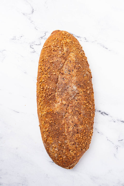 A single rustic loaf of bread with a golden-brown crust and visible flax and sunflower seeds , photographed from above on a white marble surface.