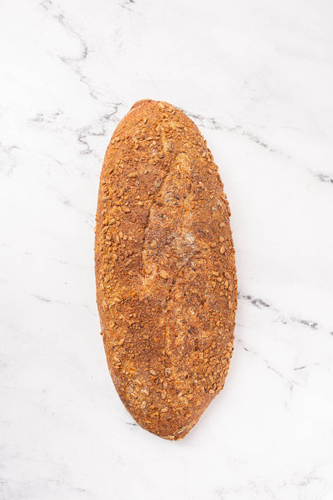 A single rustic loaf of bread with a golden-brown crust and visible seeds flax and sunflower, photographed from above on a white marble surface.