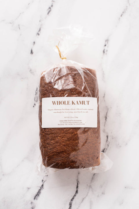 A loaf of whole kamut bread in a clear plastic bag with a white label, placed on a white marble surface.
